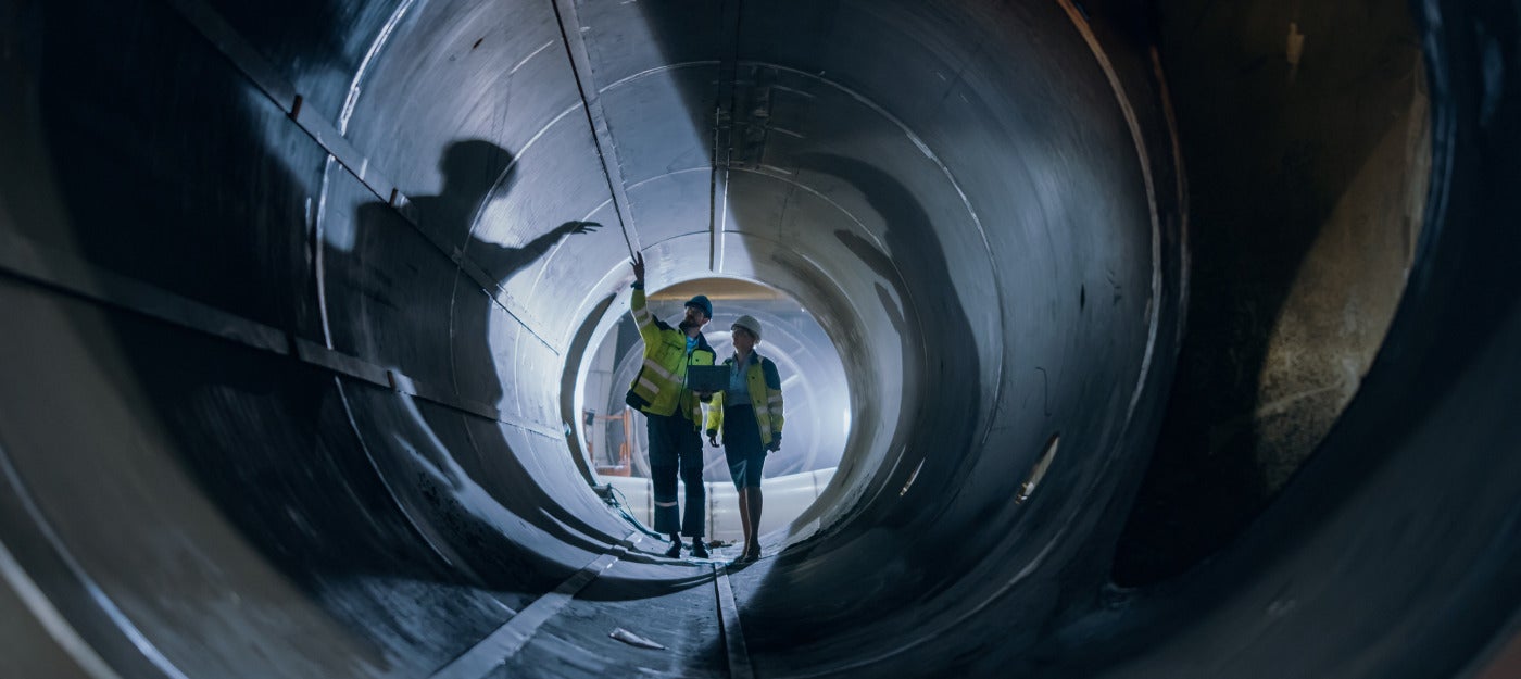 Two workers wearing safety headwear and holding a clipboard inside a pipeline microtunnel